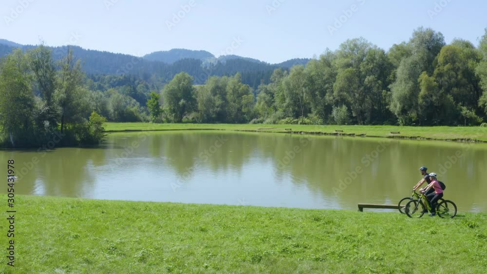 Male and female couple in their 30s riding bicycles on a lake, Aerial drone left side tracking shot, Slovenia Vodni public park all logos removed Male and female couple in their 30s riding bicycles on a lake, Aerial drone left side tracking shot, Slovenia Vodni public park all logos removed