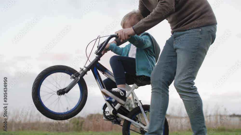 Dad helping son ride his bike supporting handlebars, having fun doing wheelies Dad helping son ride his bike supporting handlebars, having fun doing wheelies