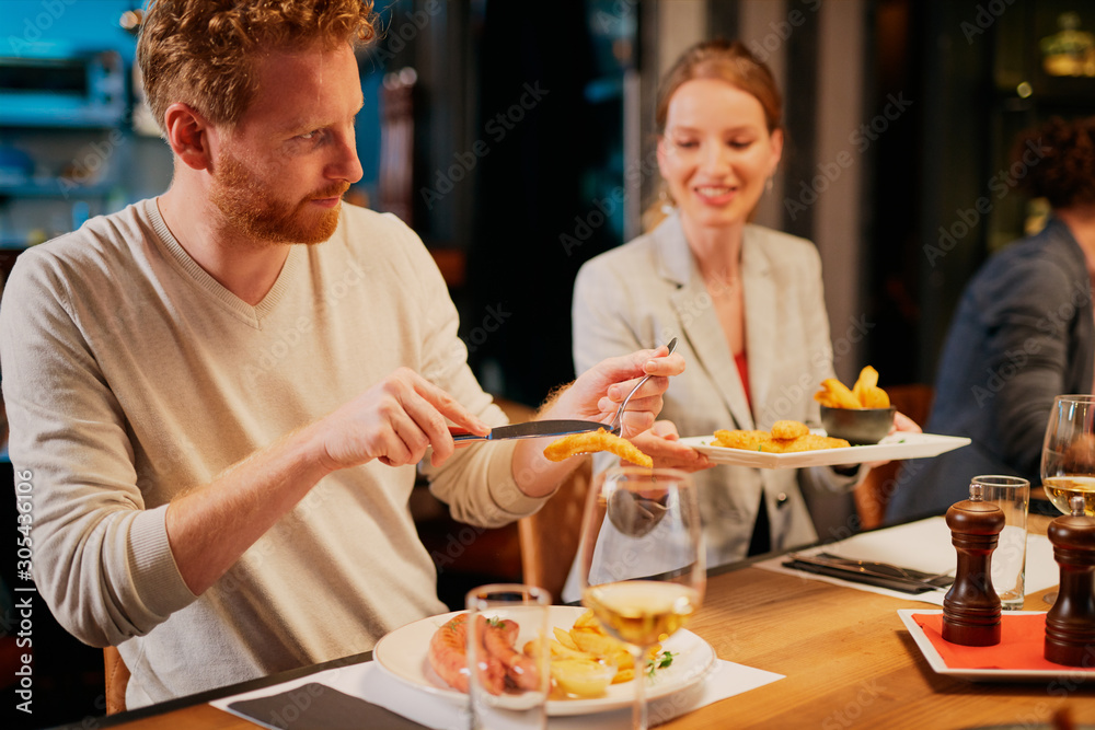 Handsome caucasian ginger taking food out of his girlfriend's plate while sitting in restaurant for dinner. In background are their friends. Handsome caucasian ginger taking food out of his girlfriend's plate while sitting in restaurant for dinner. In background are their friends.