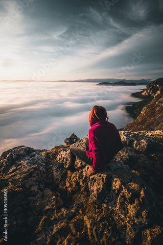 woman sitting on mountain peak watching sunrise over a sea of fog