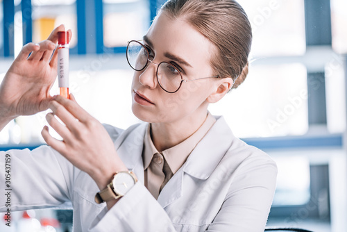 selective focus of beautiful immunologist in glasses looking at test tube with virus letters