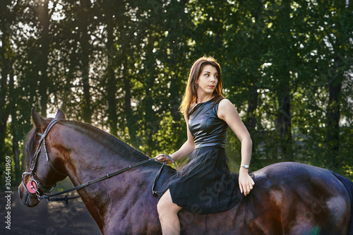 Young slim woman brunette in a black dress sitting on dark brown horse. Sunny summer evening.
