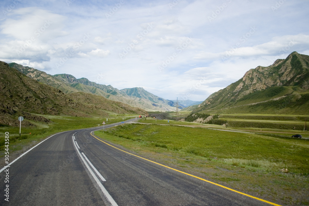 An asphalt road with a dividing strip, with road markings and signs runs along the mountains. The road is surrounded by green hills and mountains. Chuya highway in the valley of the Chuya River. Altai