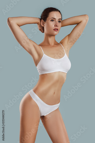 Young woman in white underwear, with bundled hair, put hands on head, posing on gray background. Plastic surgery, aesthetic cosmetology. Close-up.