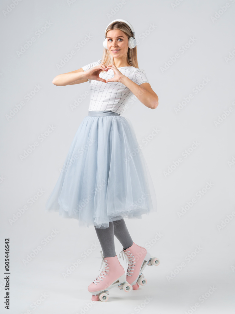 Naklejka premium Pin up woman in retro roller shoes and headphones with heart shaped hand gesture. Vertical studio shot on white background