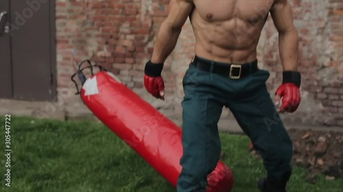 Close view. Strong handsome sportsman in red boxing gloves picking up red punching bag, throwing it to the ground over his shoulder. Slow motion