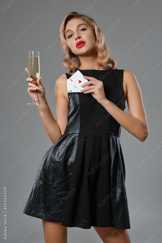 Blonde girl in black dress holding two playing cards and glass of champagne, posing against gray background. Gambling, poker, casino. Close-up. Blonde girl in black dress holding two playing cards and glass of champagne, posing against gray background. Gambling, poker, casino. Close-up.