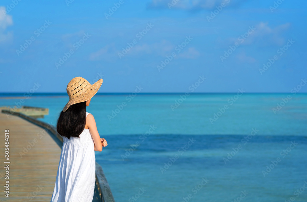 Back of young Asian girl standing on wooden bridge looking to the ocean. Teenager girl wearing sun hat or straw hat and white dress enjoy her freedom and fresh air. Concept of outdoor summer vacation. Back of young Asian girl standing on wooden bridge looking to the ocean. Teenager girl wearing sun hat or straw hat and white dress enjoy her freedom and fresh air. Concept of outdoor summer vacation.