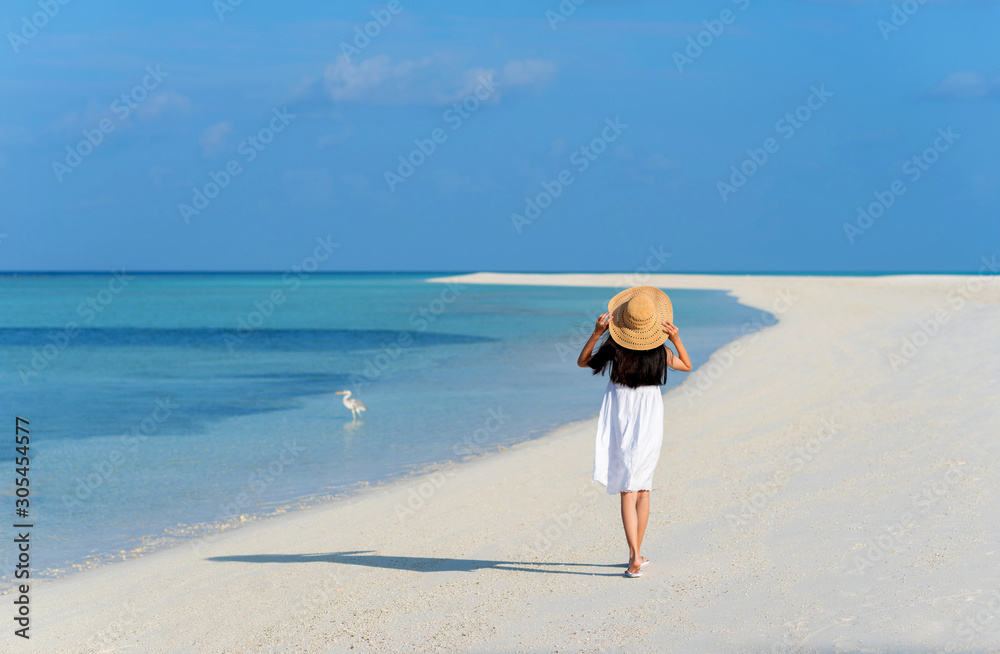 Back of young Asian girl walking on white sand beach with clear blue sea and sky. Teenager girl wearing sun straw hat, white dress enjoy vacation. Outdoor summer vacation travel concept, copy space.