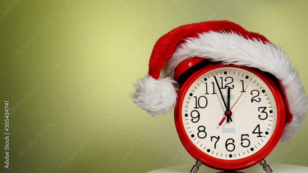Christmas time concept. Santa Claus hat and big red clock ticking showing twelve hours, on green background. Waiting for celebration. Time lapse