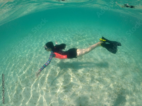 Asian teenager girl enjoy summer vacation on sunny day in crystal clear water. Half underwater of young healthy woman in colorful swimsuit, mask and black fins relaxing, free diving on turquoise sea.