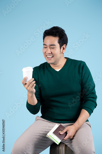 Happy Asian man sitting on lugguage, holdig map, passport and takeaway coffee mug on blue background. photo