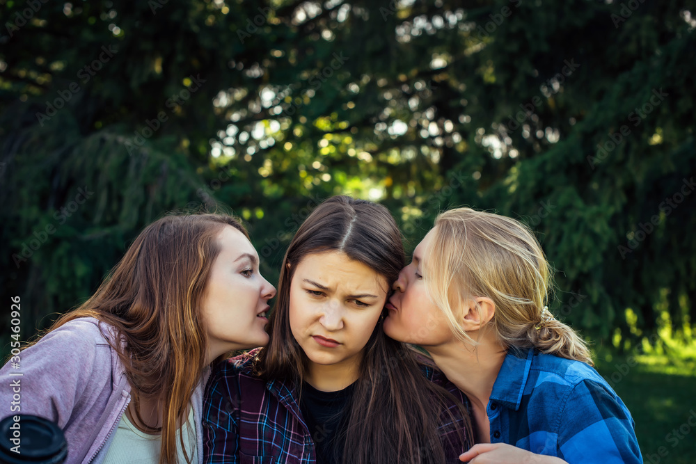 Portrait of three young girlfriends who whisper in the park. Exchange of news, emotional women whispering gossip.