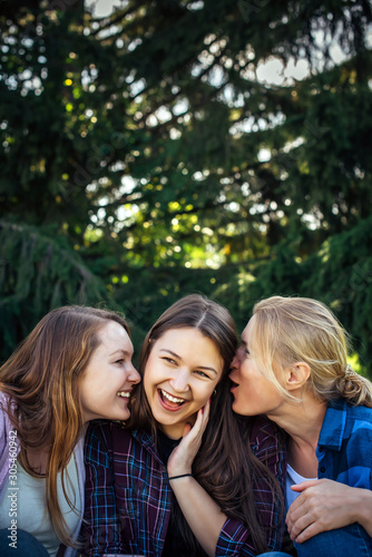 Three cheerful girls whisper and gossip against green foliage in the park. Women joke and laugh, vertical image.