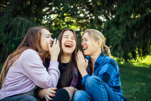 Three cheerful girls whisper and fun against green foliage in the park. Women joke and laugh, close up. Students rest outdoor.