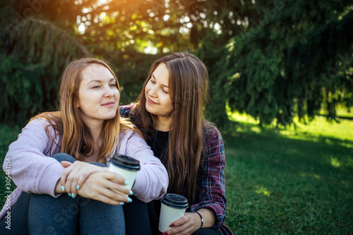 Two young beautiful girls sitting close to each other on green lawn in the park on a sunny day. Attractive women drink coffee from disposable cups outdoor. Female friendship and communication.