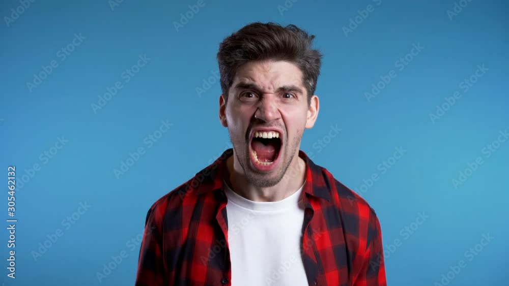 Young european stressed man in red shirt shouting isolated over blue background. Slow motion. Stressed and depressed guy loudly screaming to camera.