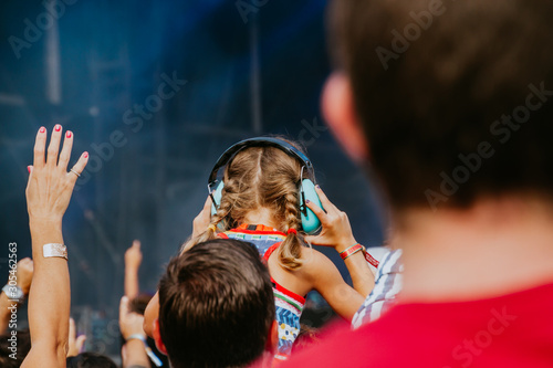 A caucasian blondie little girl wearing hearing protection helmets, at the top of some shoulders in a concert
