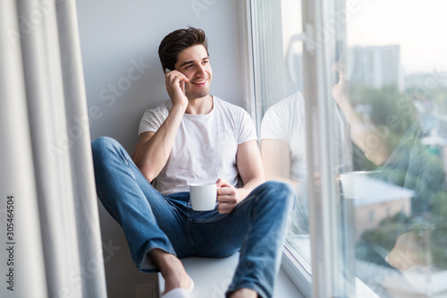 Young handsome man sitting on the windowsill and talking phone