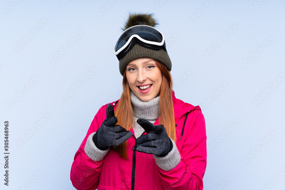 Skier redhead woman with snowboarding glasses over isolated blue wall points finger at you