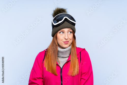 Skier redhead woman with snowboarding glasses over isolated blue wall standing and looking to the side © luismolinero