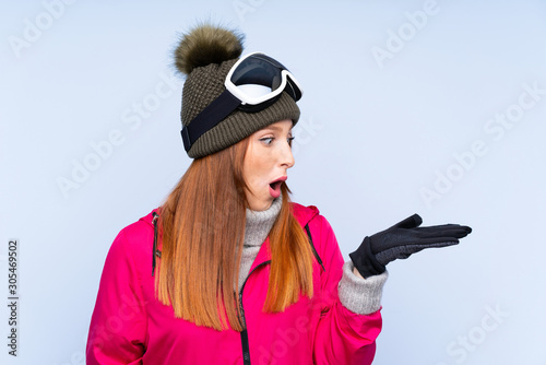 Skier redhead woman with snowboarding glasses over isolated blue wall holding copyspace imaginary on the palm