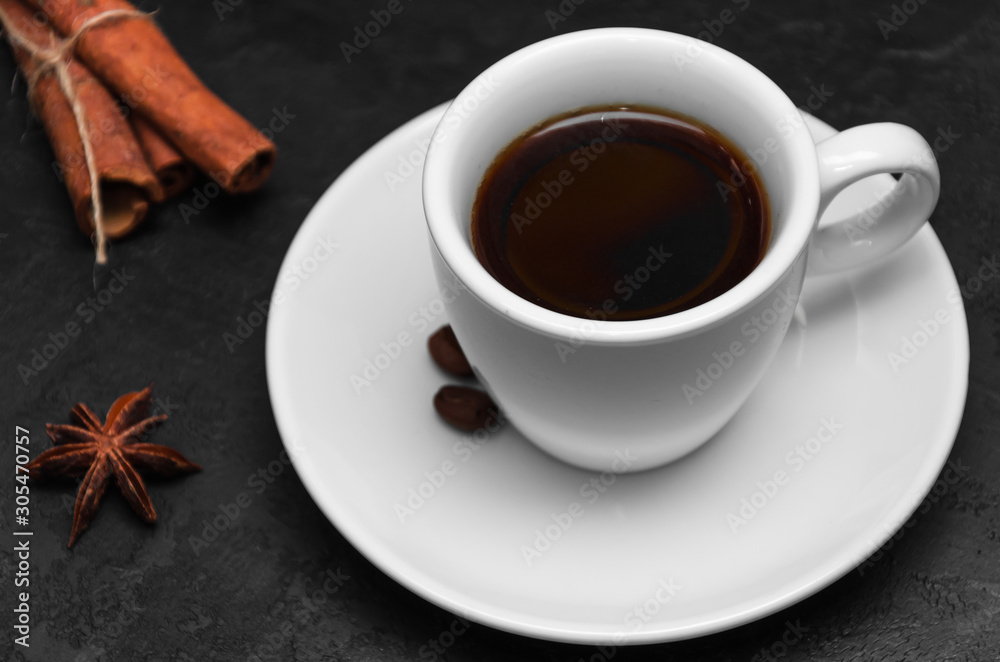 White cup of fresh hot black coffee, several coffee beans on a saucer, cinnamon sticks, anise star on black background. Coffee break concept. Closeup
