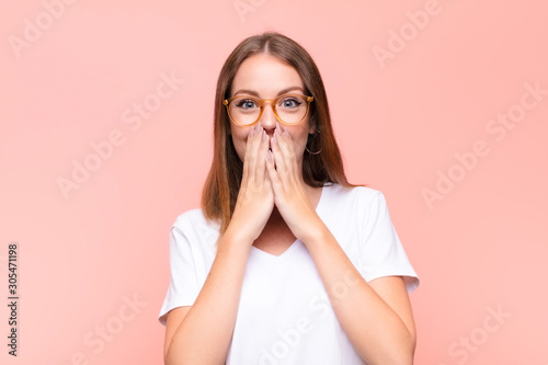 young red head woman happy and excited, surprised and amazed covering mouth with hands, giggling with a cute expression against flat wall