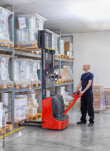 Warehouseman in storehouse moving pallet with electric stacker. Worker in warehouse stacks pallet in rack system.Caucasian labourer working with small electric forklift. Man with high lift pallet jack photo