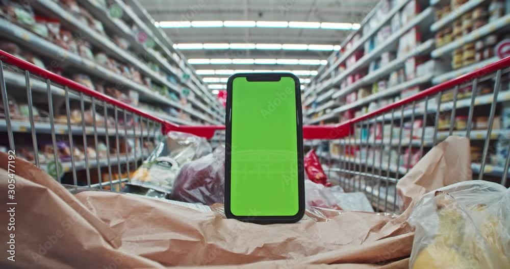 Moc-up vertical smartphone green screen with shopping list application inside cart. Close-up shopping trolley with foodstuff moving through confectionery section in supermarket.