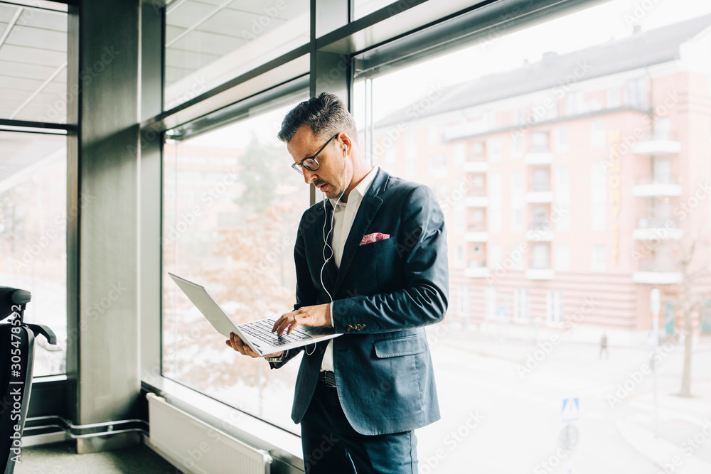 Mature businessman using laptop while standing against window in office Mature businessman using laptop while standing against window in office