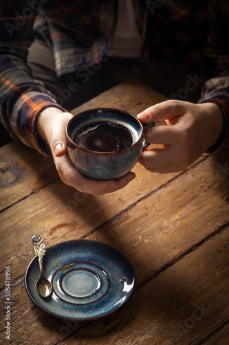 Closeup black americano coffee in blue cup in male hand, saucer with vintage tea spoon on wooden brown vintage background. Concept breakfast in coffee house