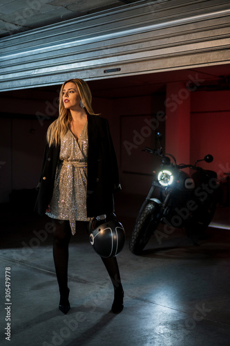 Attractive blonde woman posing for fashion photo shoot in a garage with elegant dress and a motorcycle behind