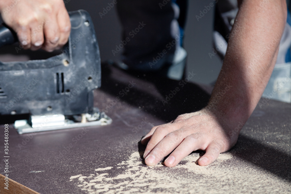 Closeup hand of woodworker with professional cutting tool fretsaw or jigsaw, cut wooden tabletop, sawing plank, brown filings, sawdust. Concept professional installation and repair Closeup hand of woodworker with professional cutting tool fretsaw or jigsaw, cut wooden tabletop, sawing plank, brown filings, sawdust. Concept professional installation and repair