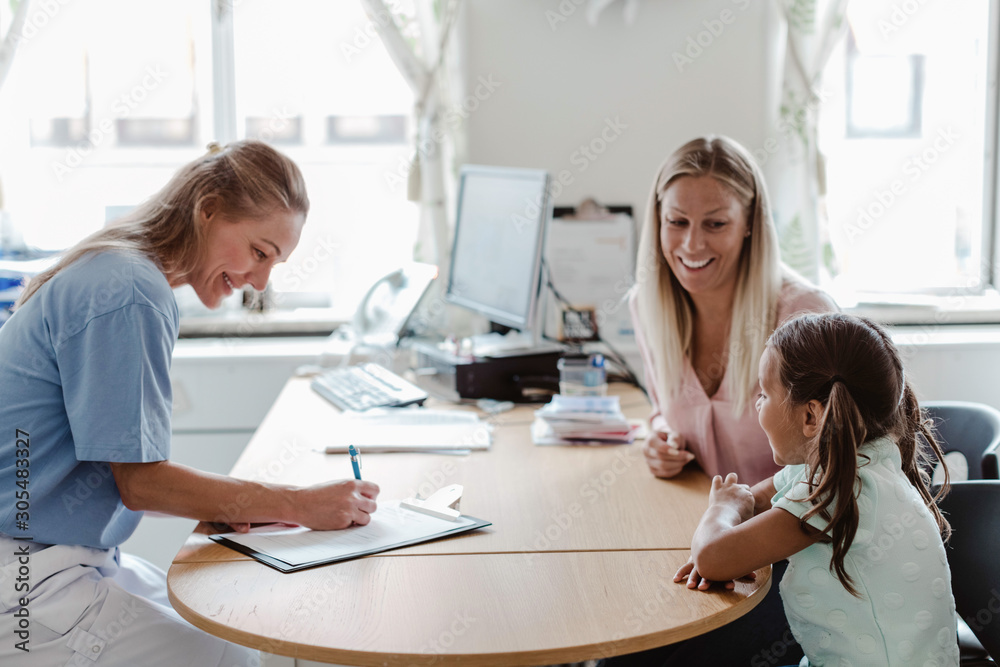 Smiling pediatrician writing prescription while girl sitting with mother at desk in clinic Smiling pediatrician writing prescription while girl sitting with mother at desk in clinic