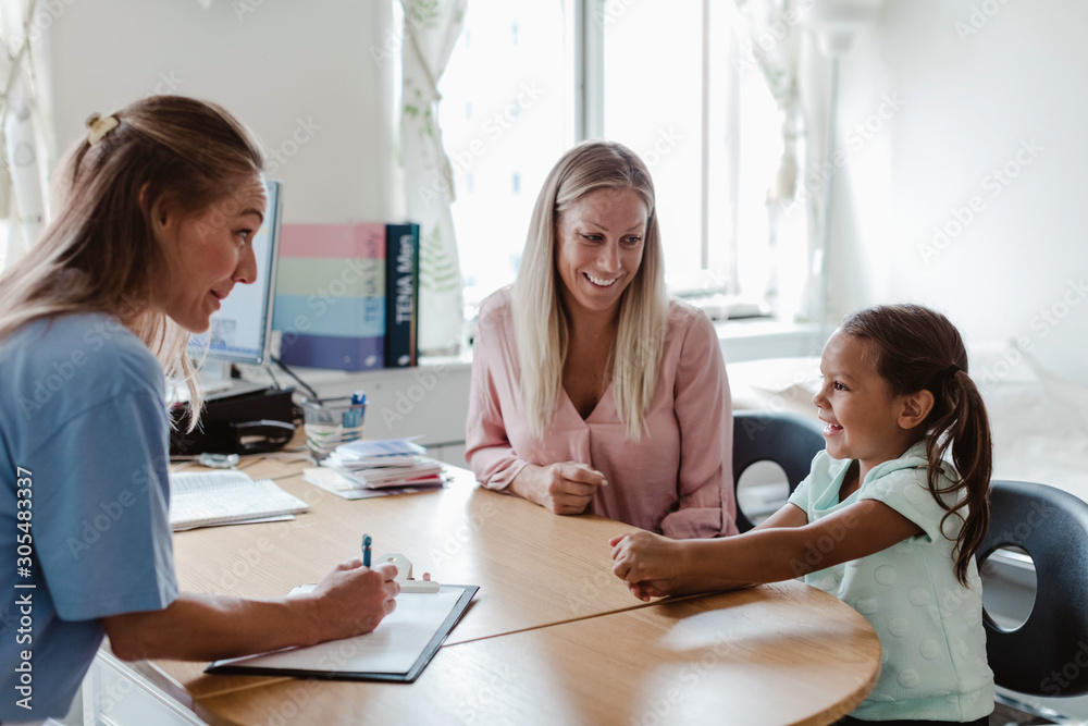 Female pediatrician and mother looking at smiling girl sitting at desk in hospital Female pediatrician and mother looking at smiling girl sitting at desk in hospital