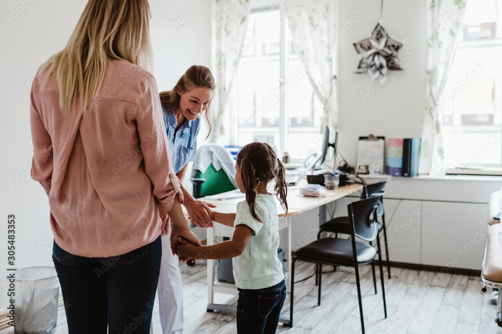 Smiling pediatrician shaking hands with girl while mother standing in doctor's office