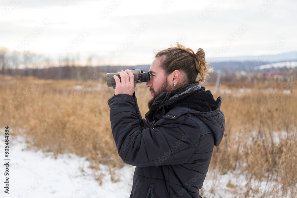 Medium horizontal side view of young bearded man in dark jacket and hair tied back staring into field glasses during a early winter grey afternoon, Quebec City, Quebec, Canada