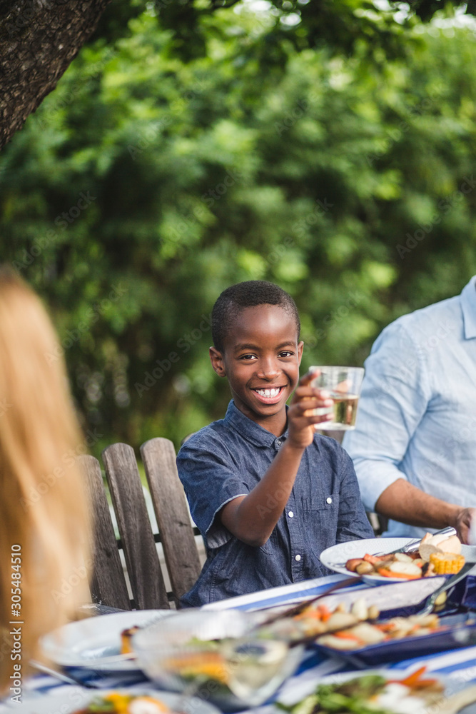 Smiling boy holding drinking glass while sitting with family in backyard during party Smiling boy holding drinking glass while sitting with family in backyard during party