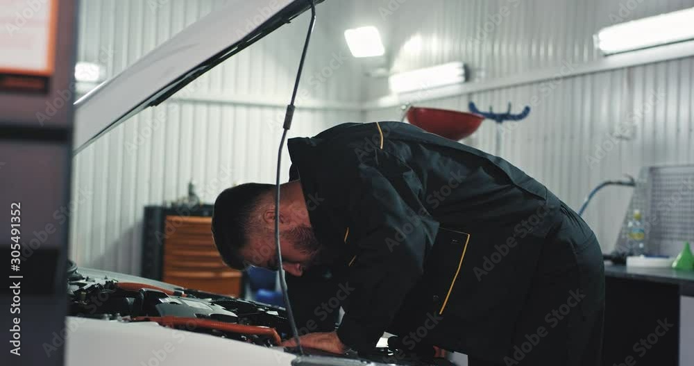 Mechanic young guy in a uniform concentrated working on a car fixing the problem he get sweating Mechanic young guy in a uniform concentrated working on a car fixing the problem he get sweating