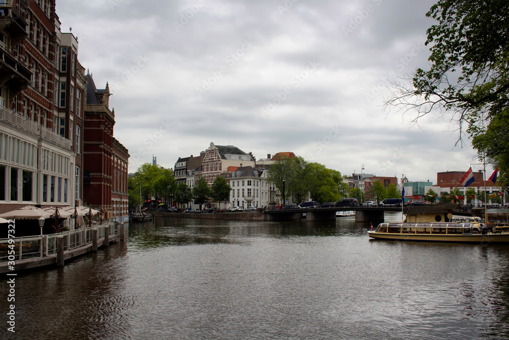 View of canal, boats, historical and traditional buildings in Amsterdam. It is a summer day with cloudy sky.