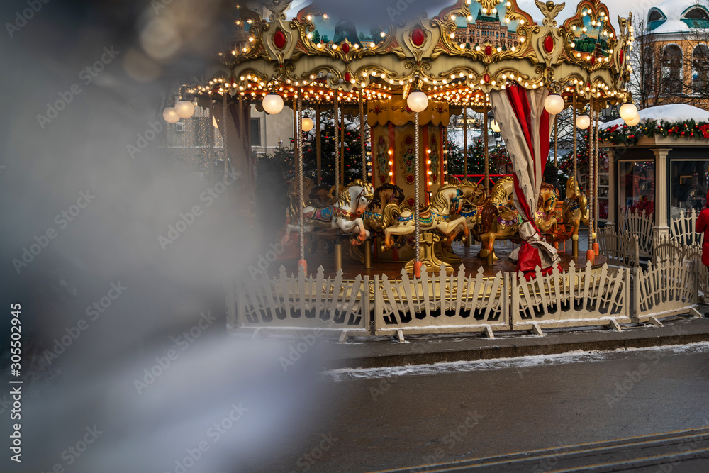 Obraz premium Сarousel horses to traditional Christmas market. Outdoor fair on winter day. View through the frost pine branch. Happy new year. Festive street decor in winter holidays.
