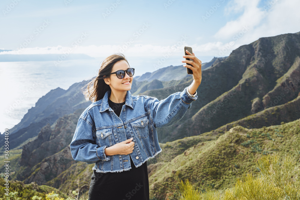 Young woman traveler taking a selfie using a smartphone on the background of the ocean and mountains. Young woman traveler taking a selfie using a smartphone on the background of the ocean and mountains.