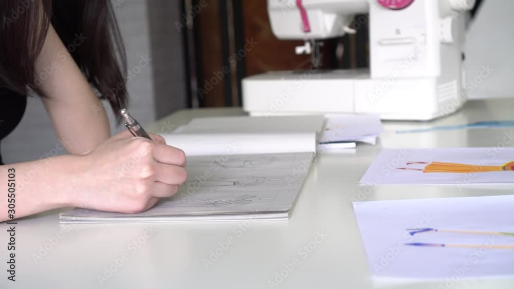 Happy adult Asian fashion designer focusing and creating new project in notebook while sitting at desk with sewing tools against blurred workshop interior