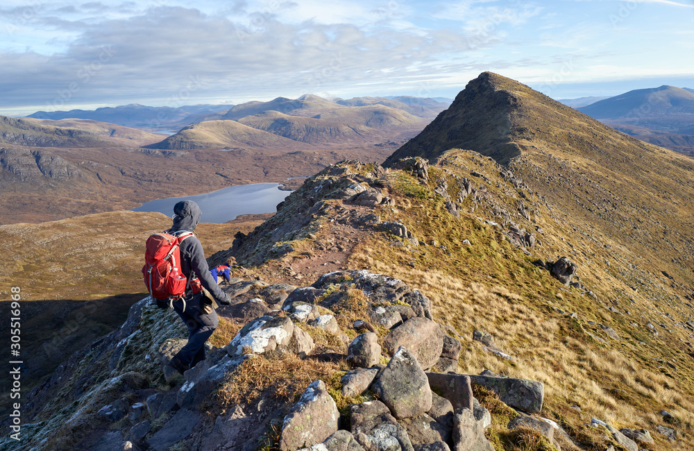 A hiker and their dog walking over the narrow ridge that leads to Sgurr an Tuill Bhain with Lochan Fada in the distance in the Scottish Highlands.