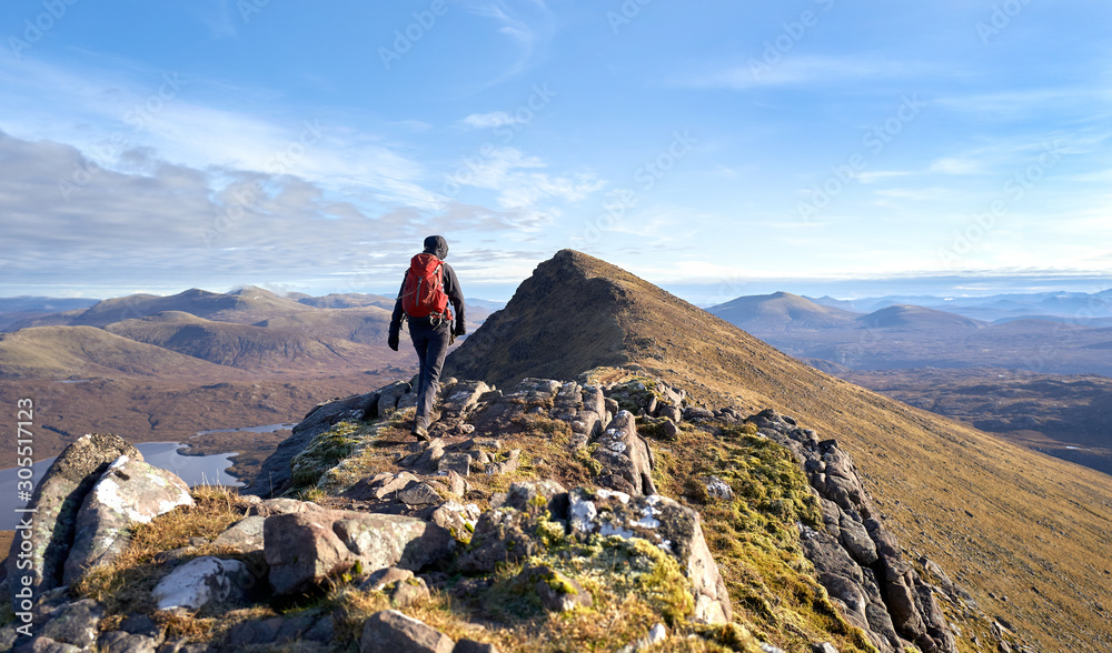 A hiker walking towards the summit of Sgurr an Tuill Bhain along a narrow rocky ridge in the Scottish Highlands on a sunny winters day.
