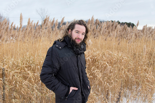 Medium horizontal view of young bearded man in dark jacket and long hair tied back turning and staring in a field during a early winter grey windy afternoon, Quebec City, Quebec, Canada © Anne Richard