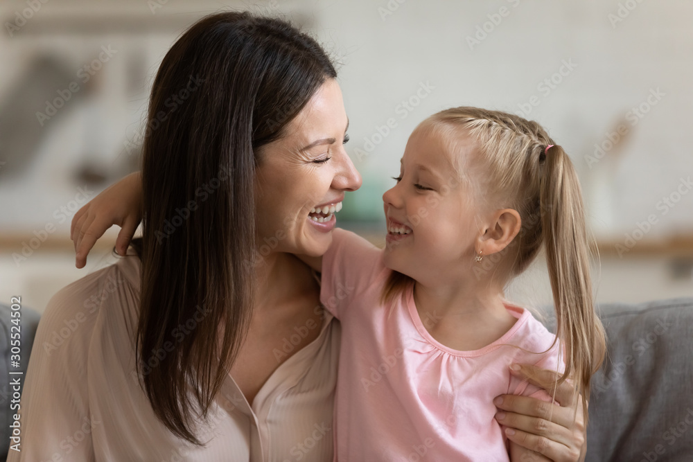 Cheerful mom embracing adorable little daughter laughing sit on sofa