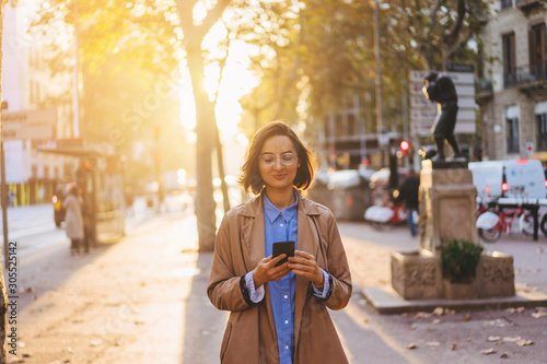 Positive young woman dressed in casual wear installing app on mobile phone while smiling at camera, portrait of hipster girl holding smartphone device standing in outdoors and using public 4G internet