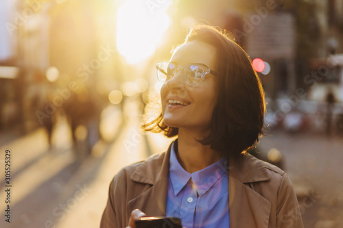 Happy millennial hipster girl in glasses laughing on urban setting during sunny day, positive cheerful woman tourist dressed in casual look holding smartphone gadget in hand for communicate
